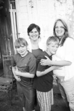 picture of co owners Erica and Deana with arms around their sons. Standing in front of a white door.