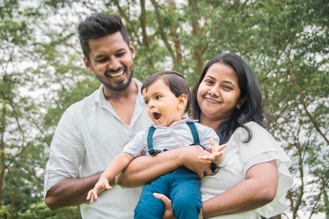 Family of three standing outdoors with their baby, trees in the background